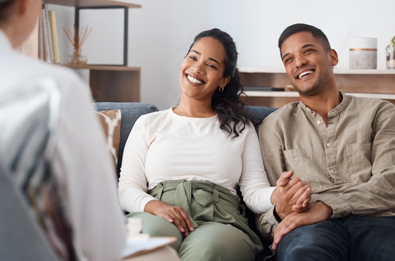 Happy couple on sofa, therapy and marriage counselling, support and love with relationship advice together. Smile, man and woman with therapist in conversation, healthy communication and healing help AdobeStock_606466908